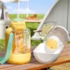 6fc352a4-9ac8-456b-84e3-fcbe7796989c.jpg Outdoor product photo: yellow baby bottle with teal hip strap beside a white bottle warmer on a wooden table; dog in the blurred background